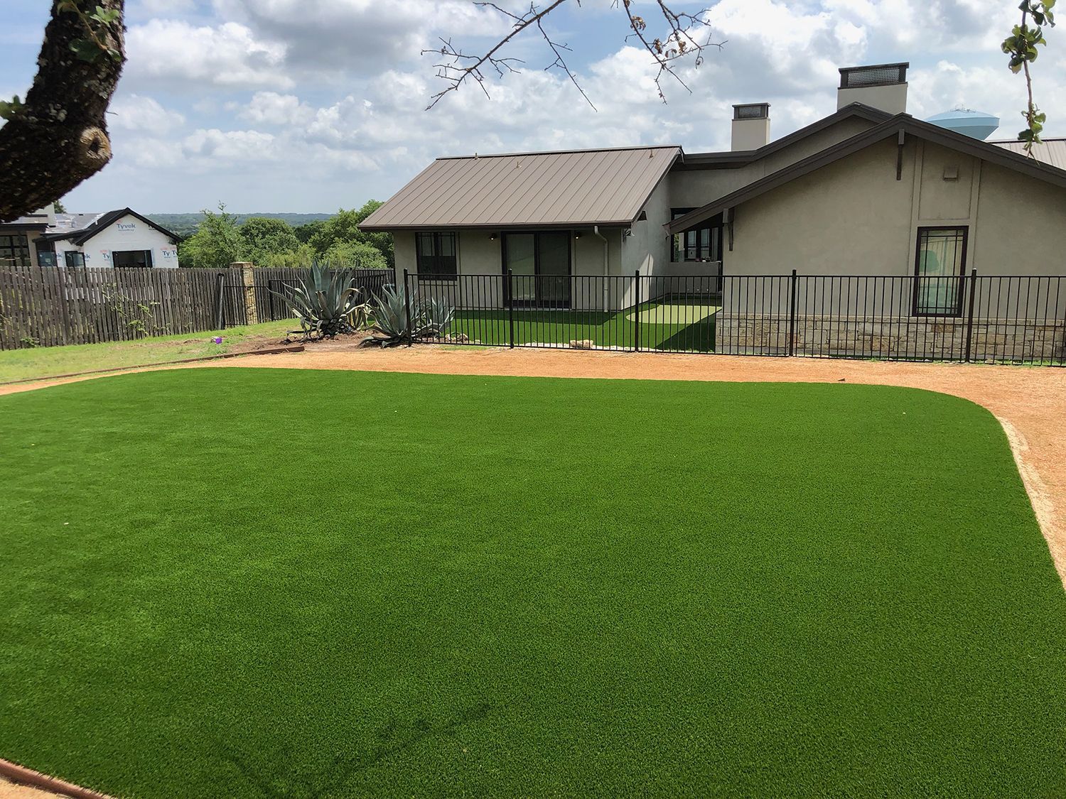 Backyard with green turf, tan walkway, fence, and a house on a sunny day.