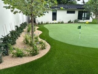 A putting green with a curving border, landscaping, and a white house in the background.