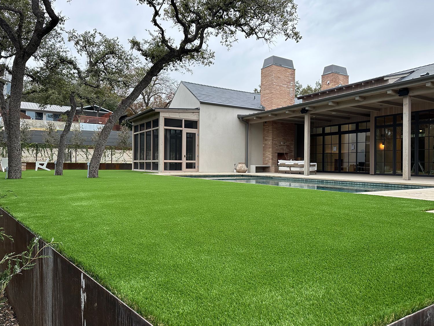 Backyard with green lawn, pool, and house. Features trees and a screened-in porch under an overcast sky.
