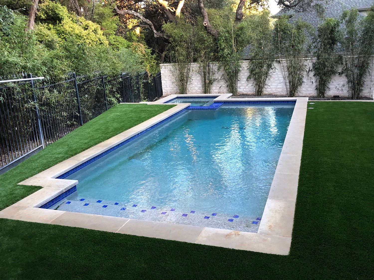 Rectangular swimming pool with a stone border and turf lawn. Small hot tub at the end, surrounded by greenery.