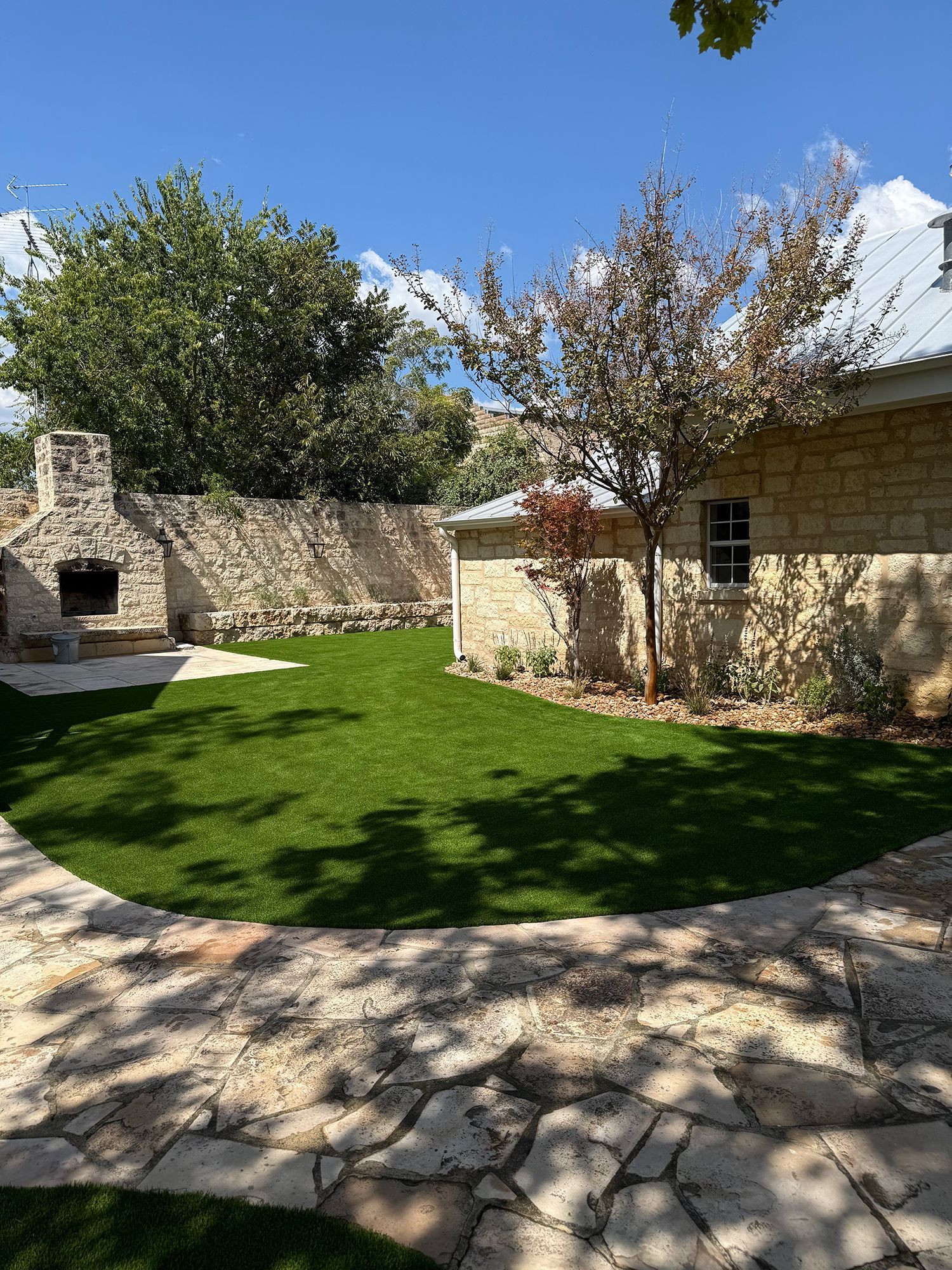 Backyard with green lawn, stone patio, stone building, and outdoor fireplace under a blue sky.