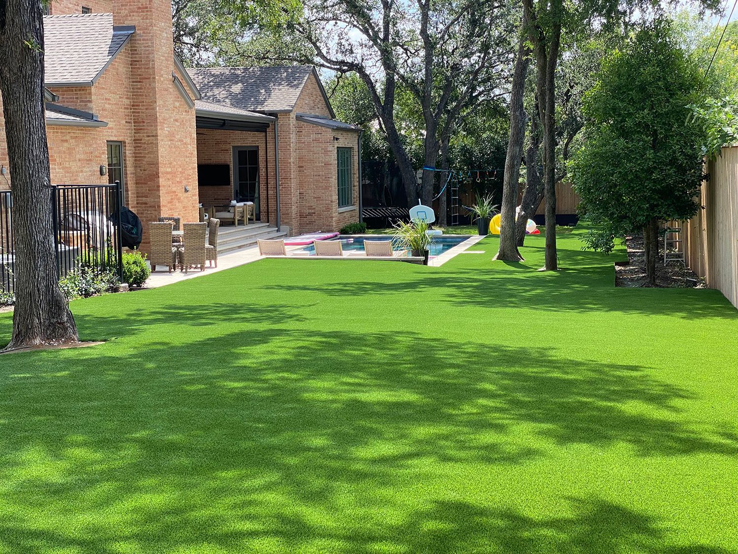 A backyard with a brick house, swimming pool, green lawn, and trees.