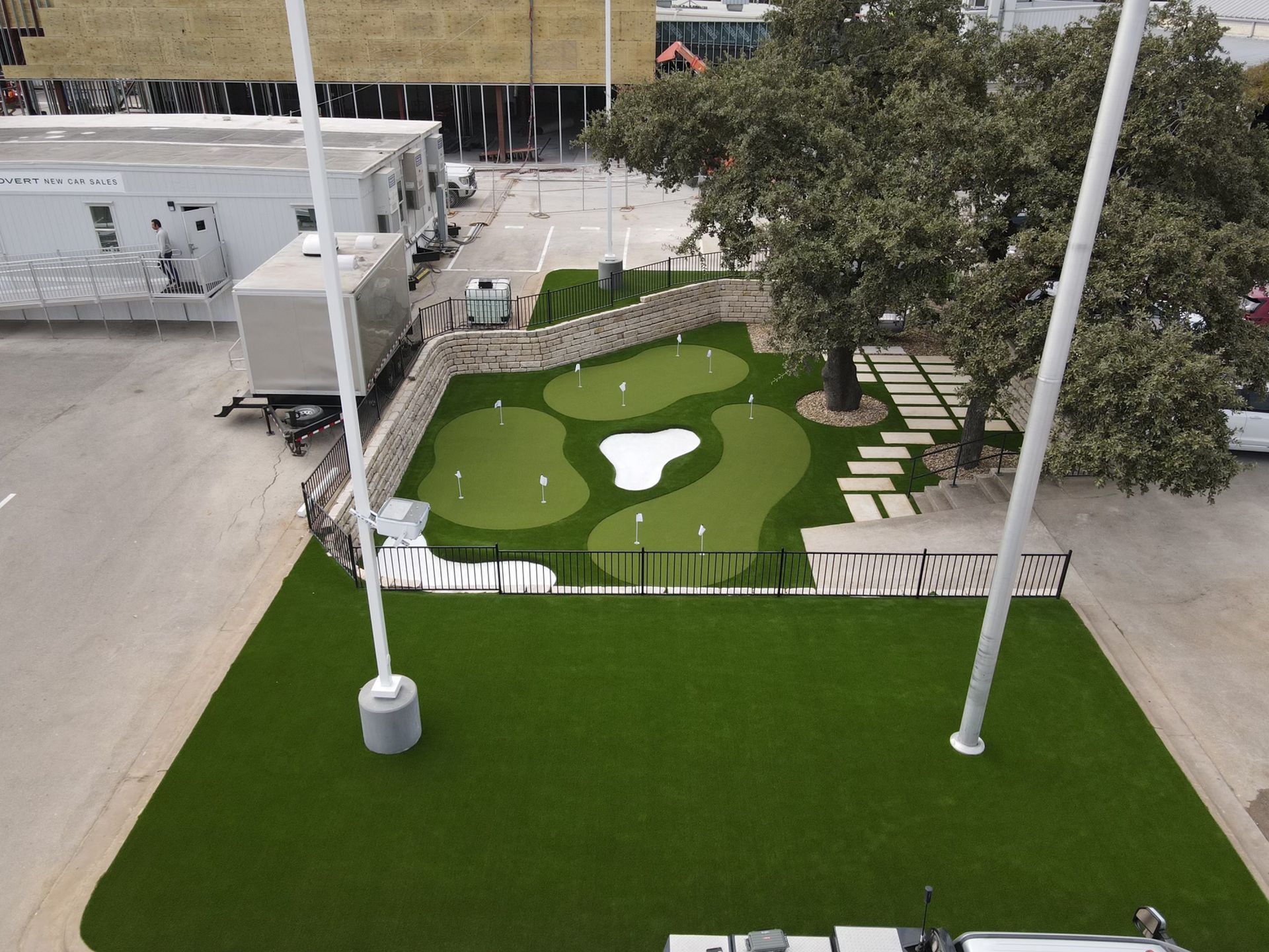 Aerial view of a putting green with several holes, surrounded by landscaping, trees, and fencing.