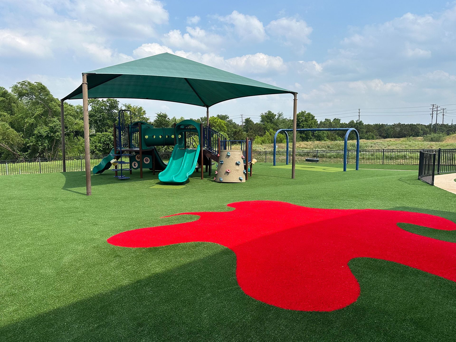 Playground with a green shade, slides, swings, and red splash pad. Green grass and blue sky.