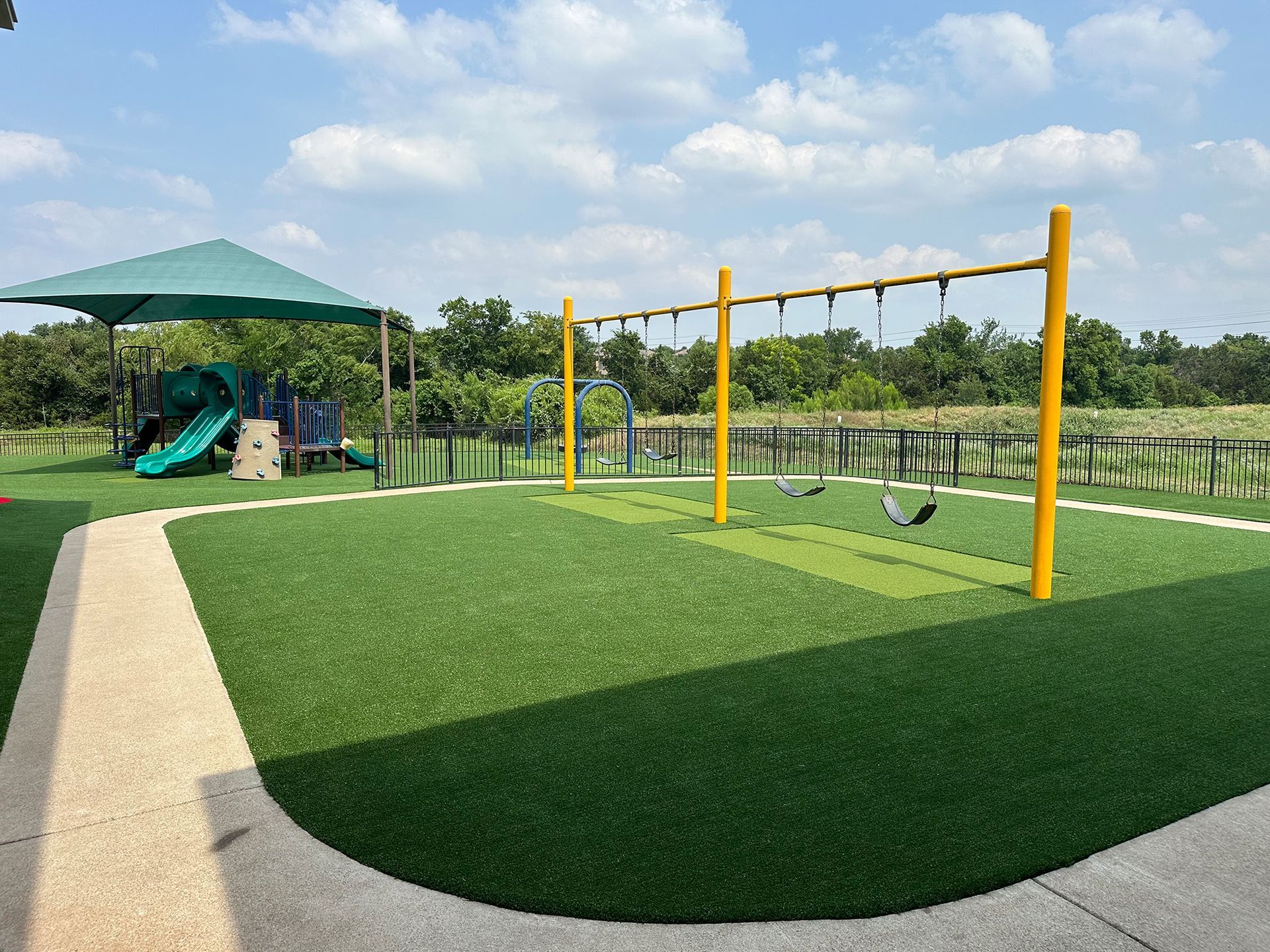 Playground with yellow swings, green turf, and shade structure under a blue sky.