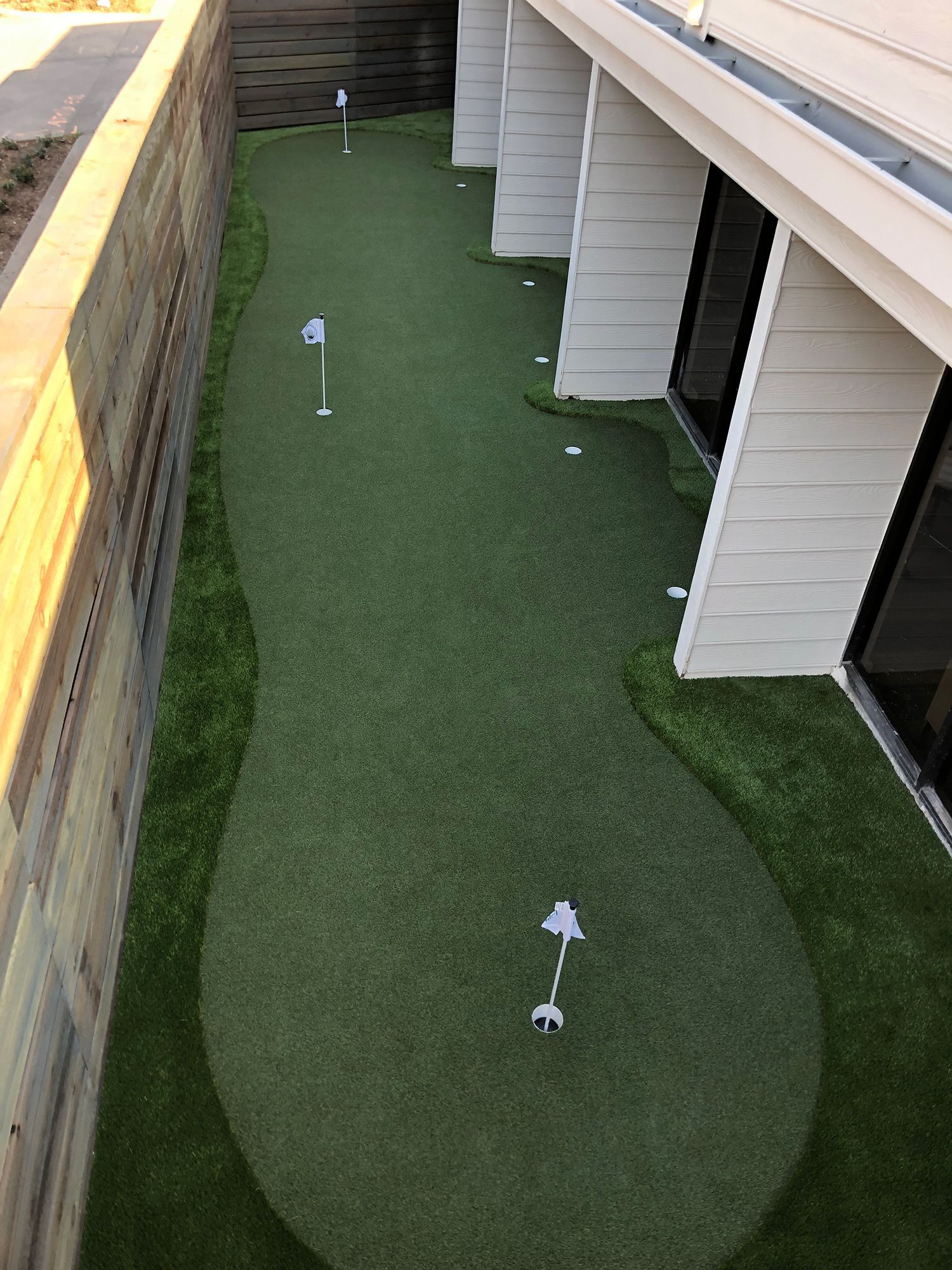 Artificial turf putting green between a wooden fence and building with flagsticks.