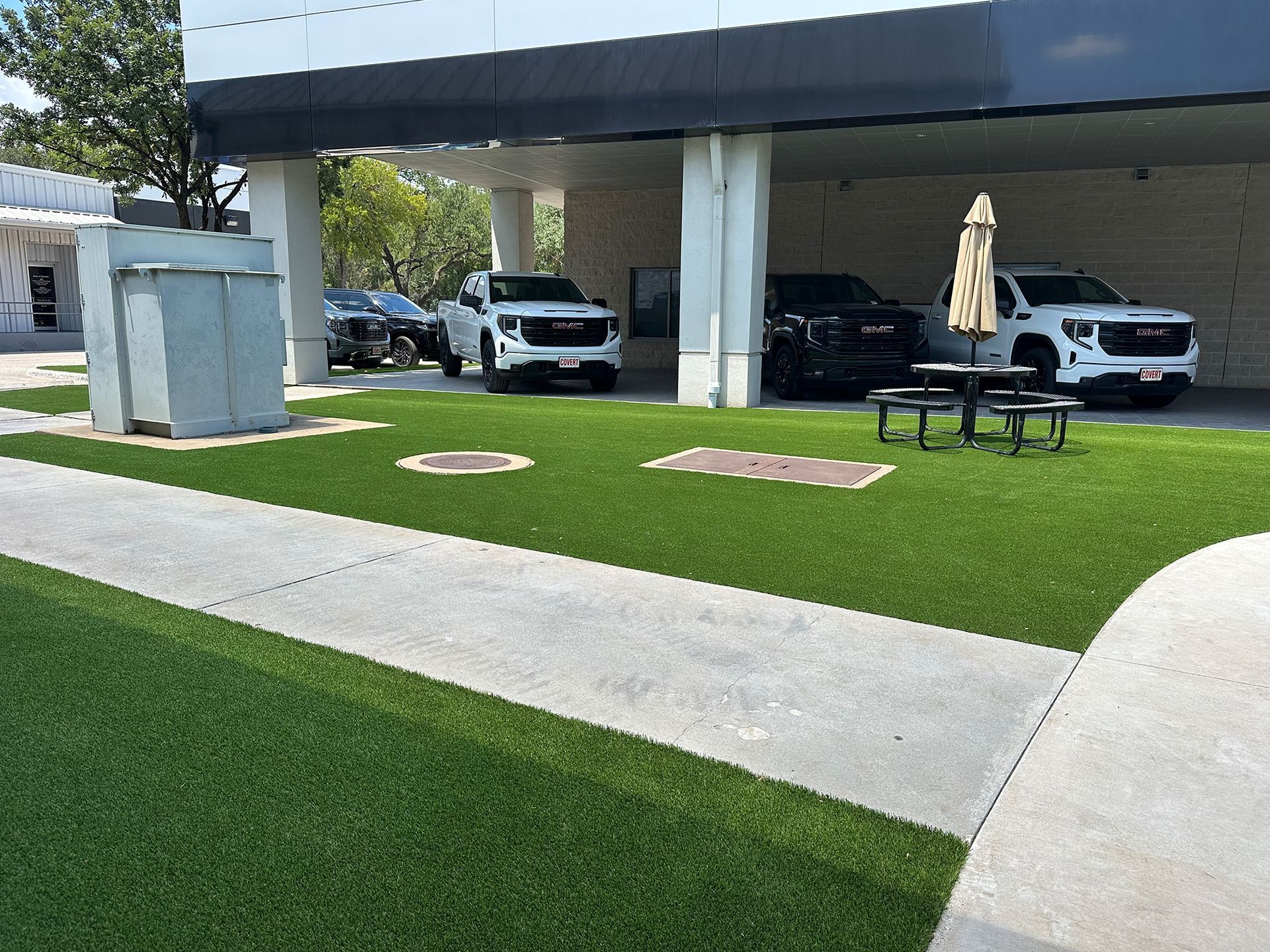 Cars parked under a building's awning on artificial turf; picnic table and umbrella nearby.
