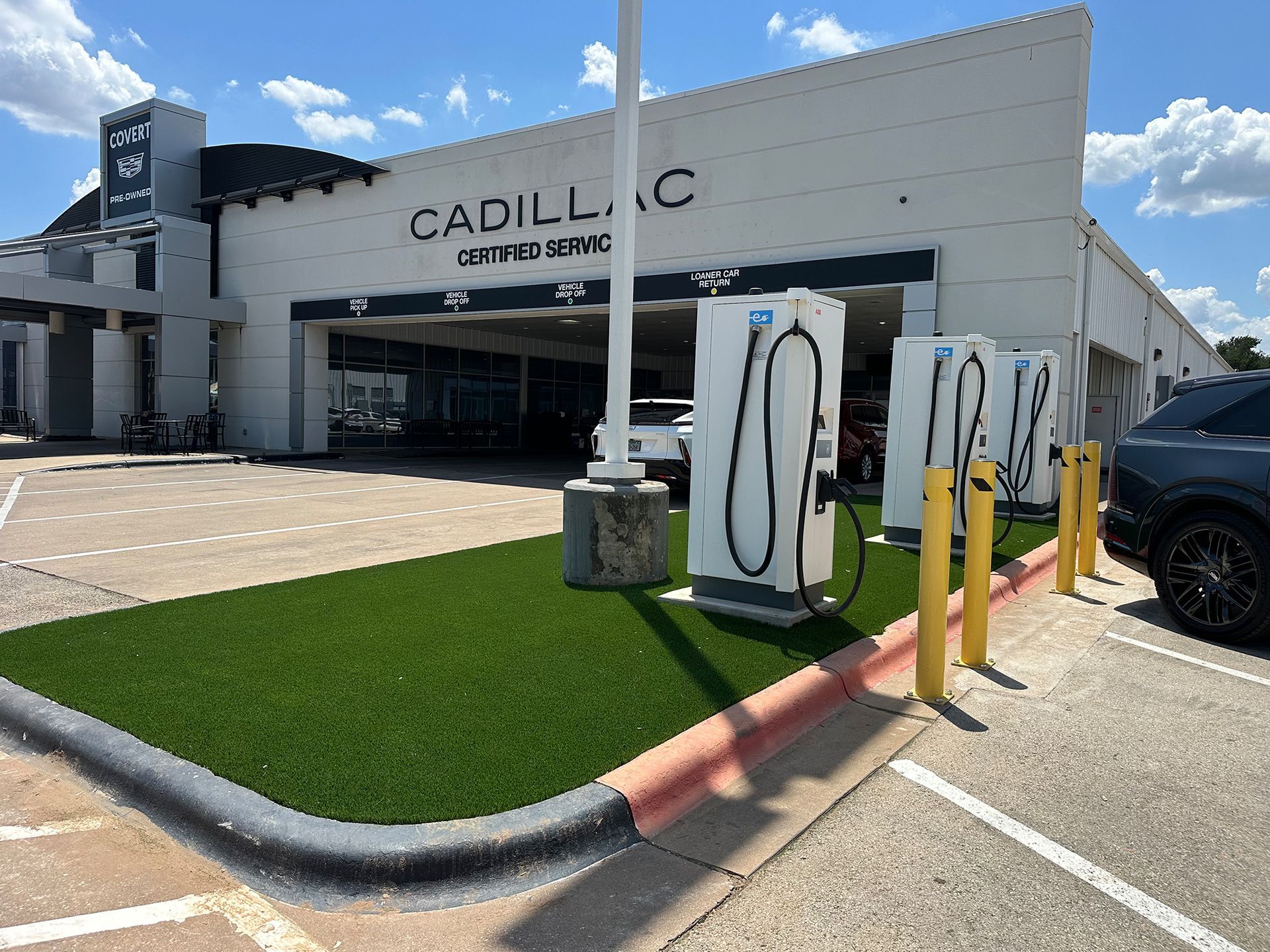 Cadillac dealership with electric vehicle charging stations in front. White building with artificial turf.