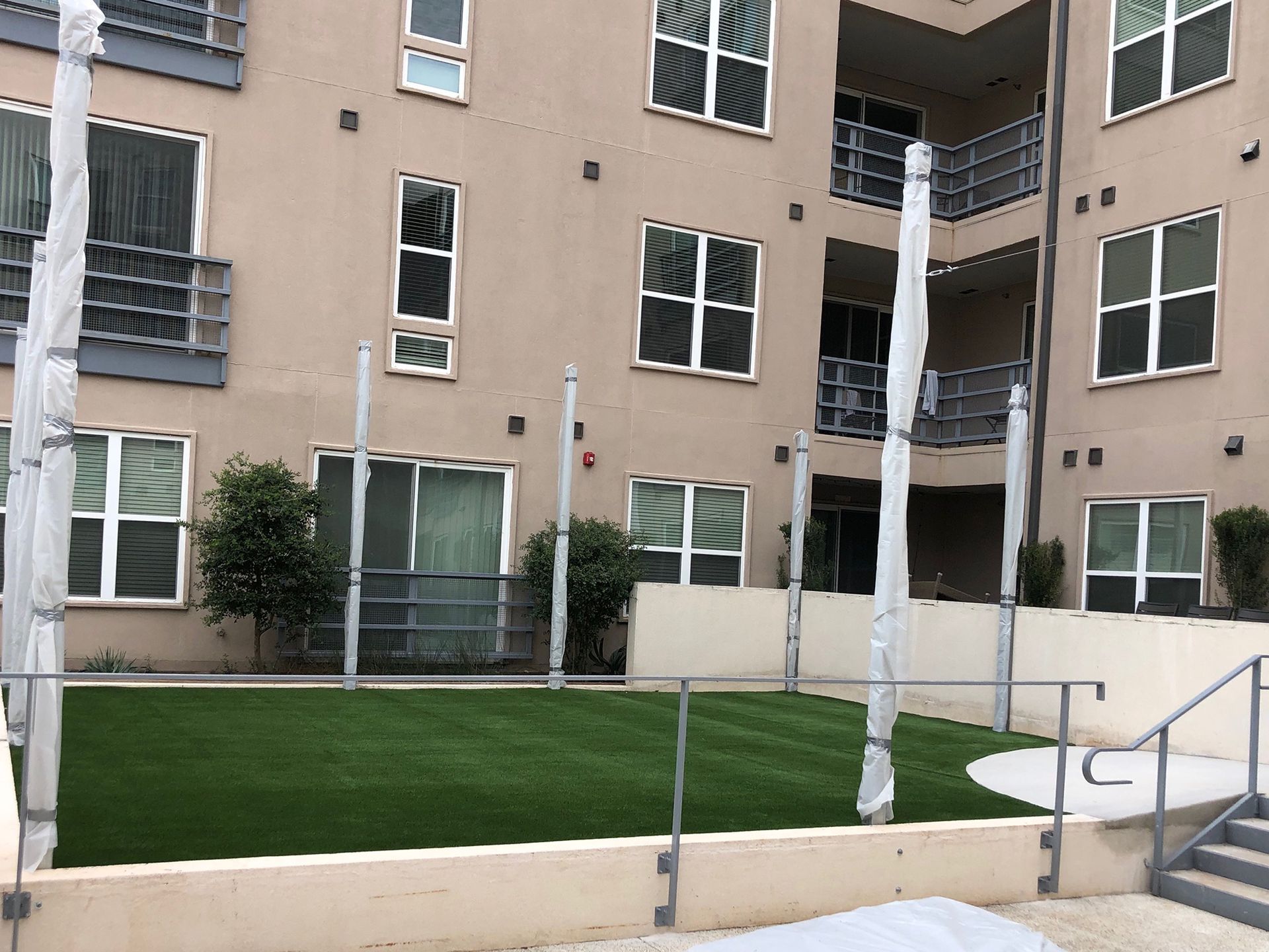 Courtyard with artificial turf, surrounded by building, railings, and poles wrapped in plastic.