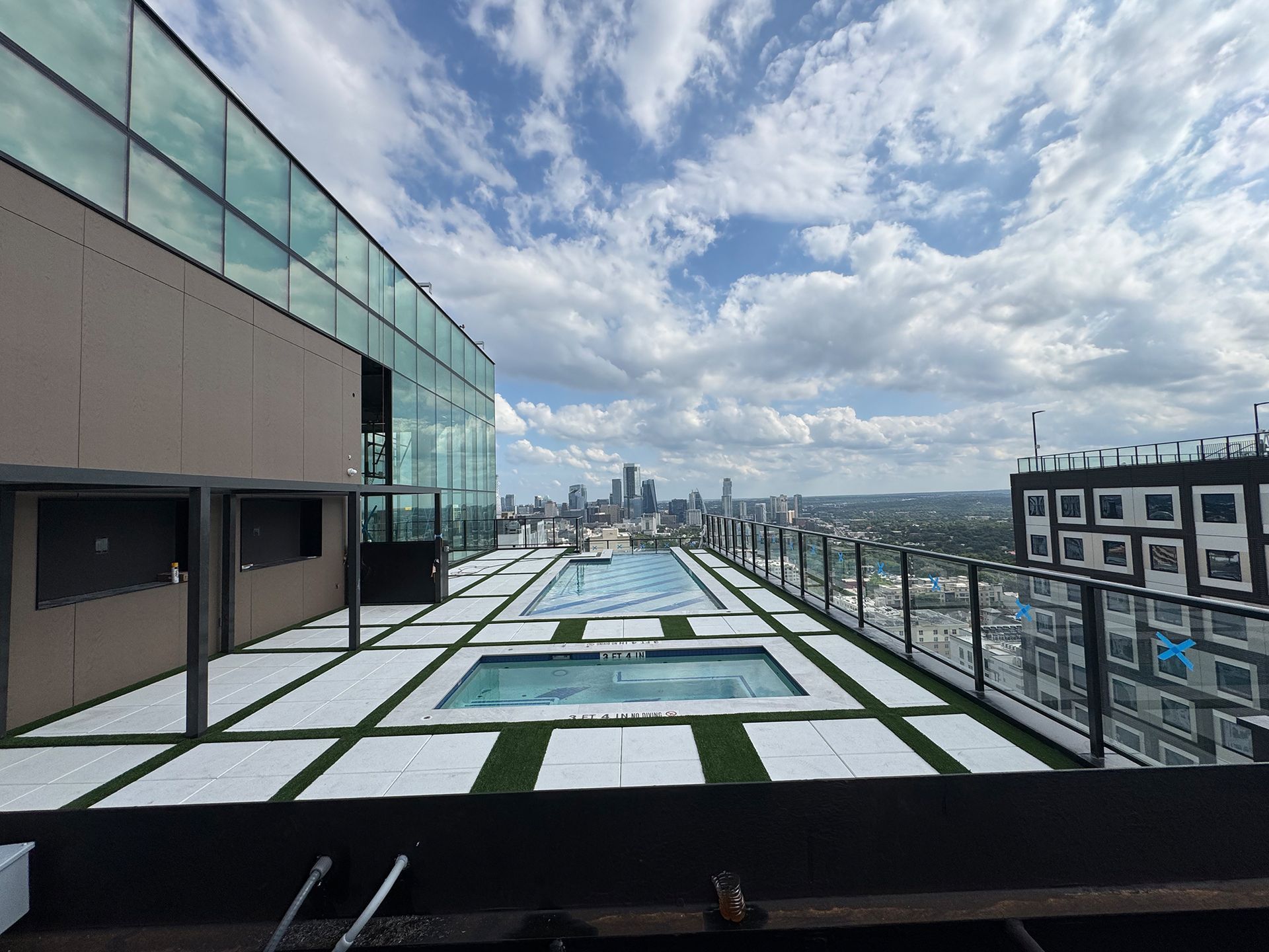 Rooftop pool deck with city skyline view, surrounded by glass and metal railings. Sunny day with clouds.