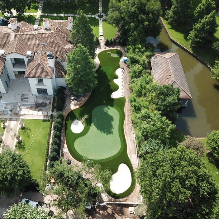 Aerial view of a backyard putting green with sand traps, next to a large house and boathouse.