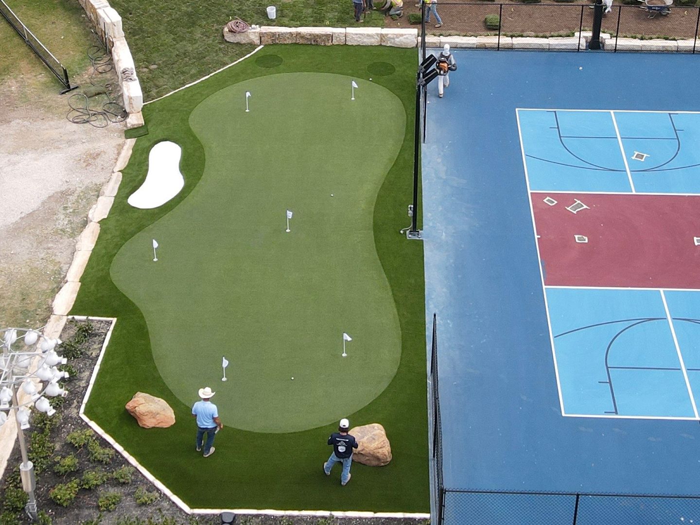 Overhead view of a putting green and basketball court. Two people play golf near the green.