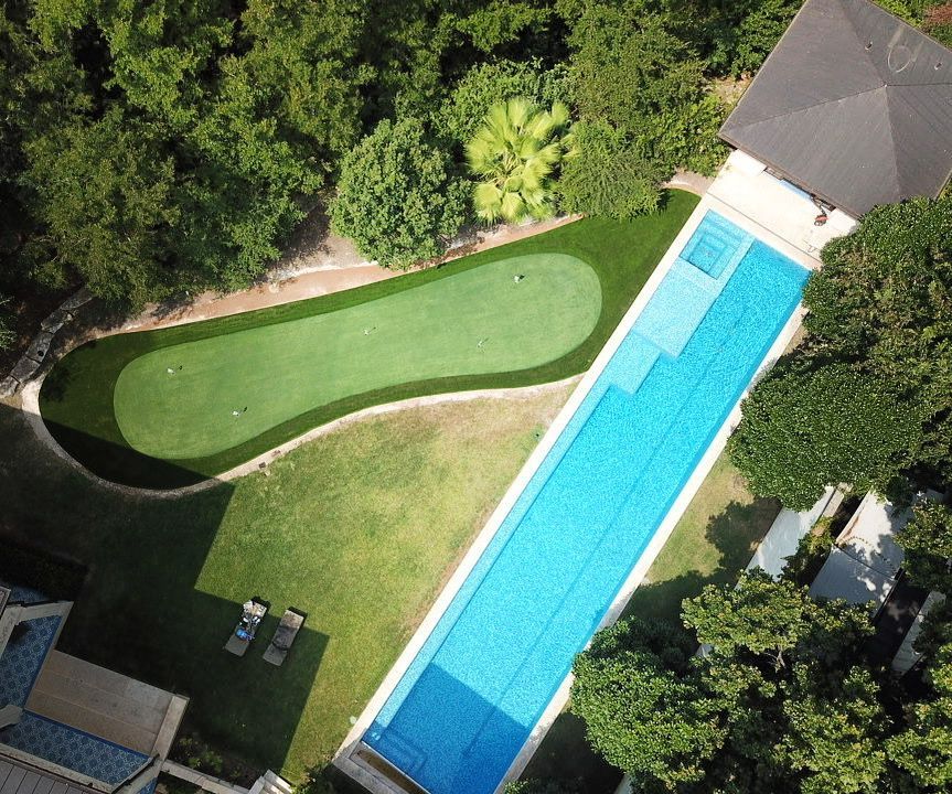 Aerial view of a long swimming pool, putting green, and lush green trees. Sunny outdoor setting.