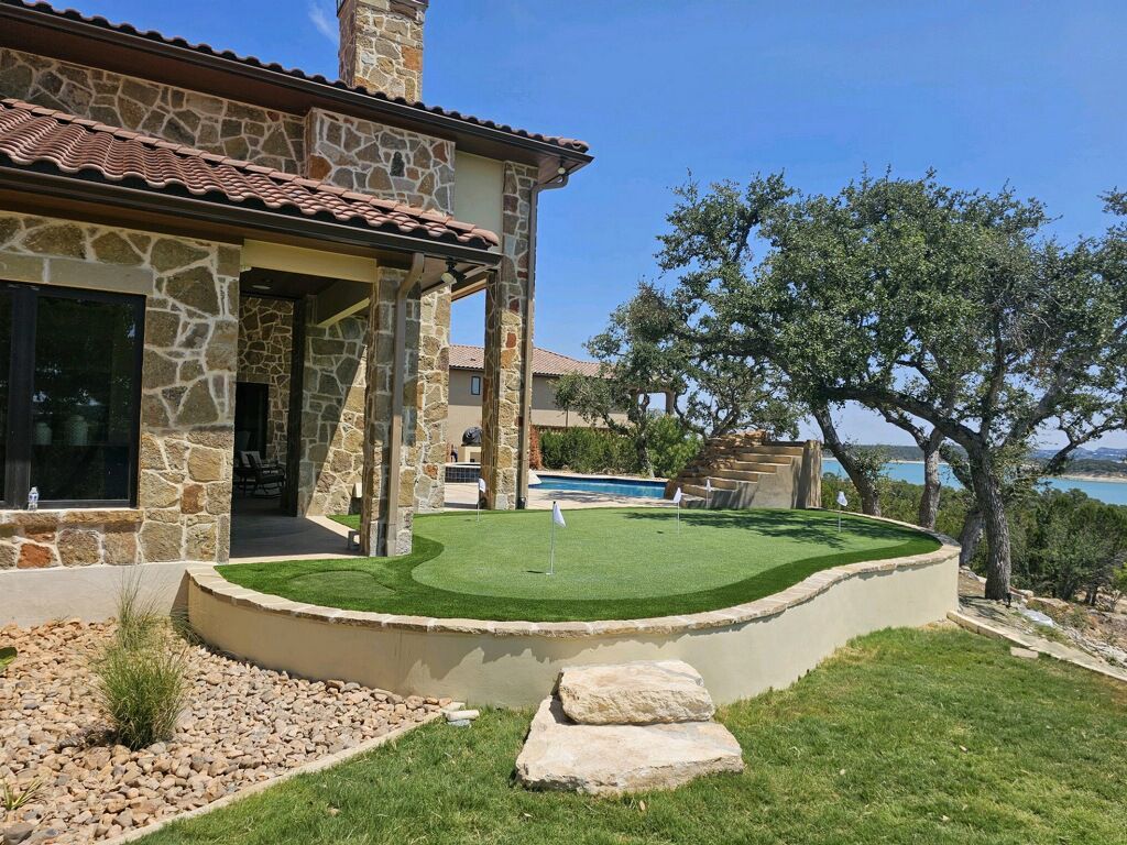 Backyard with stone house, putting green, pool, and lake view on a sunny day.