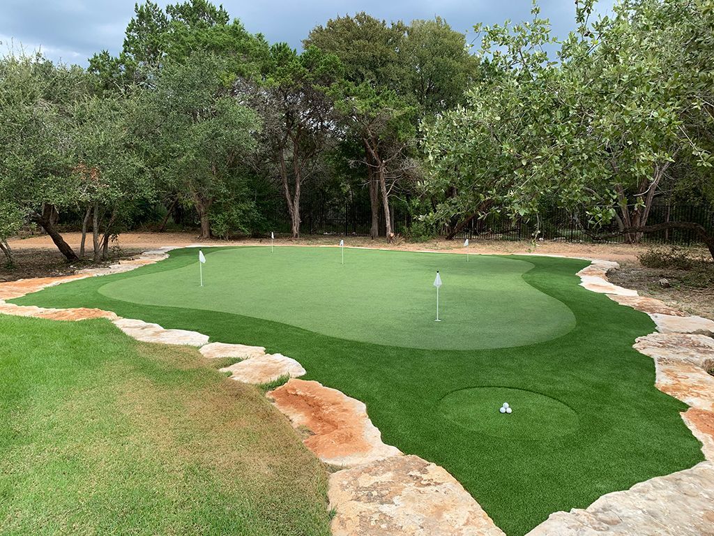 Green artificial turf putting green with rock border, surrounded by trees.
