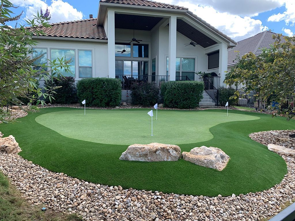 Backyard putting green with flags, rocks, and house in the background. Green grass, blue sky.