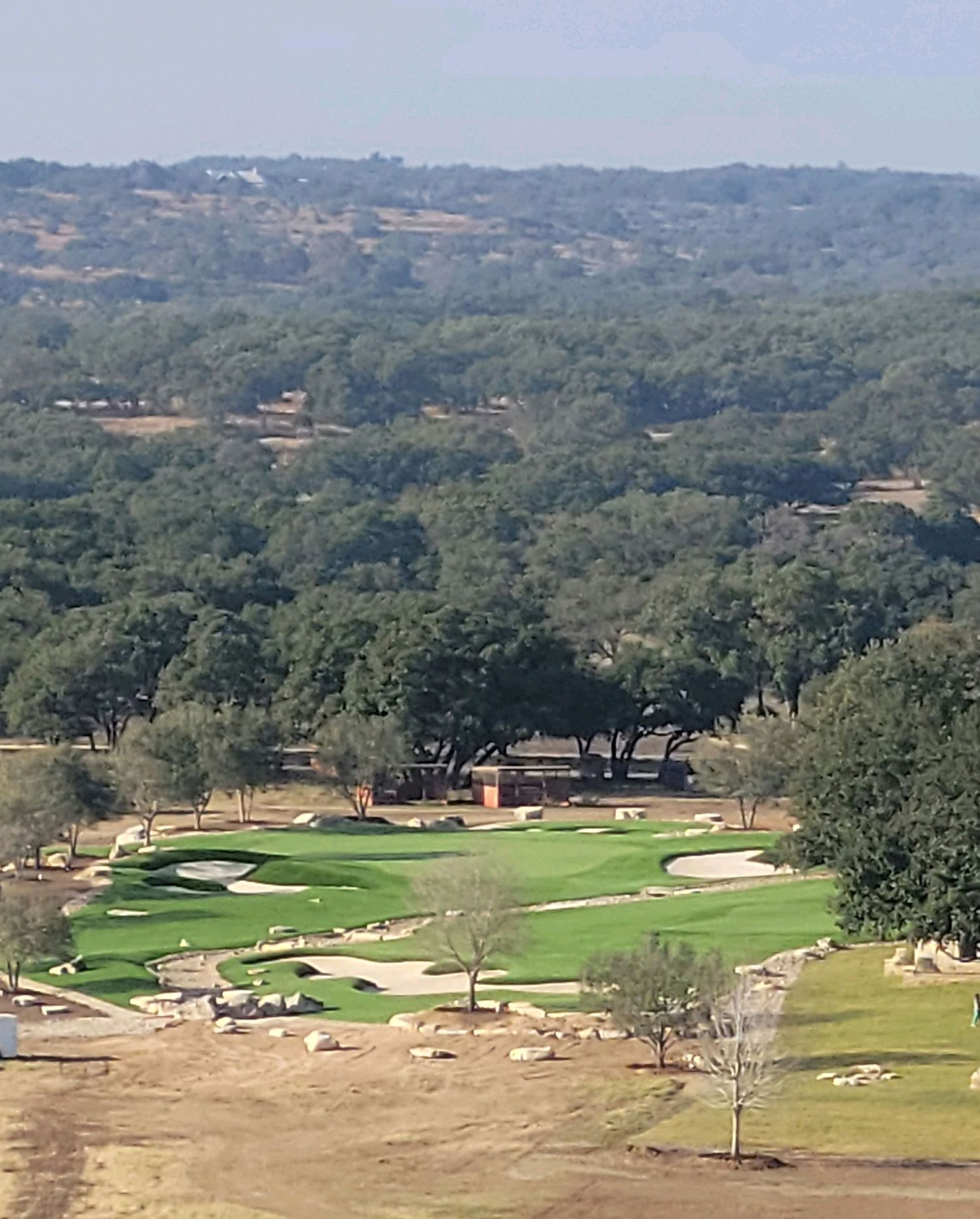 Golf course with green fairways, sand traps, and trees set in a valley with distant hills.
