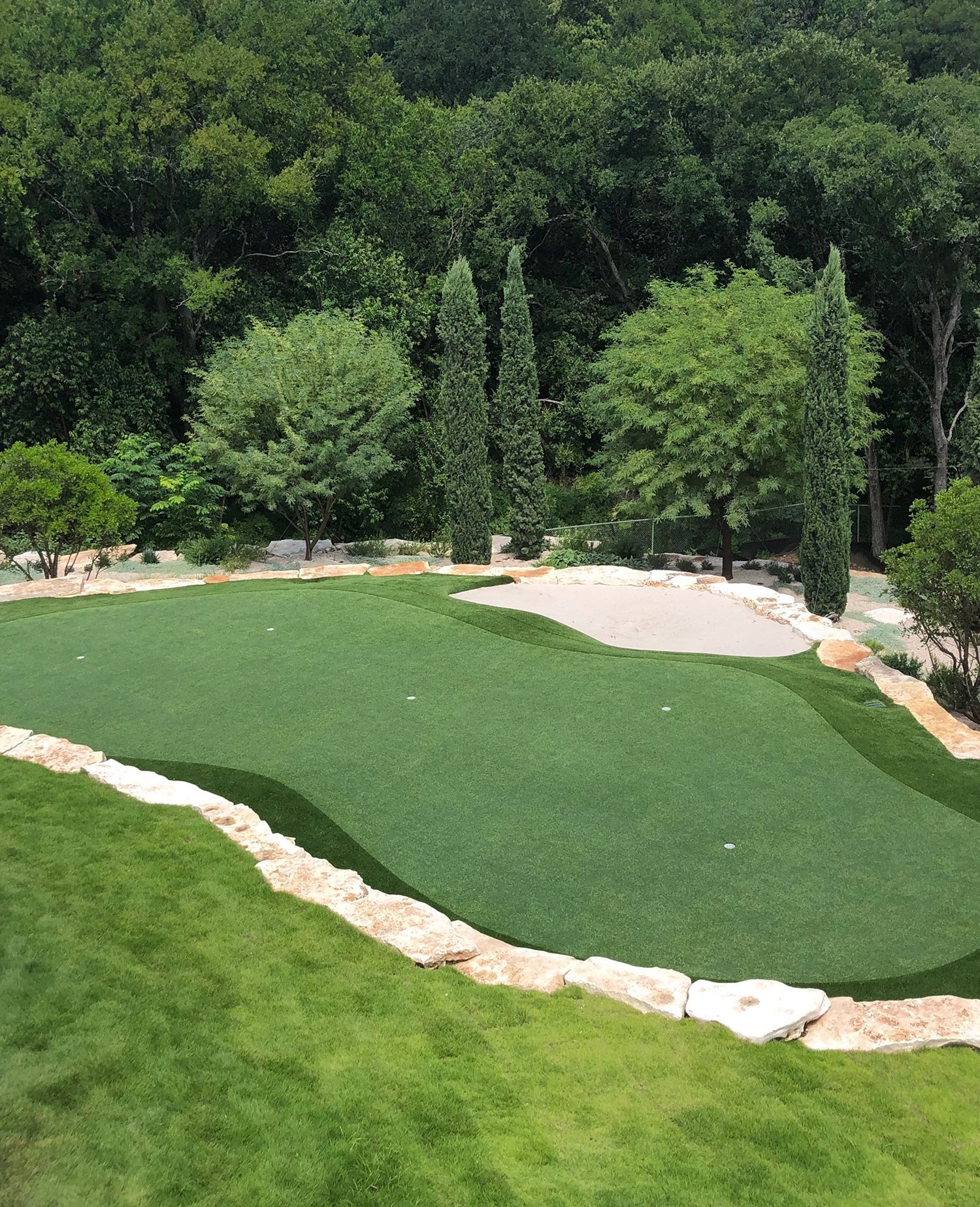 Green artificial turf putting green with sand trap, surrounded by trees and stone border.