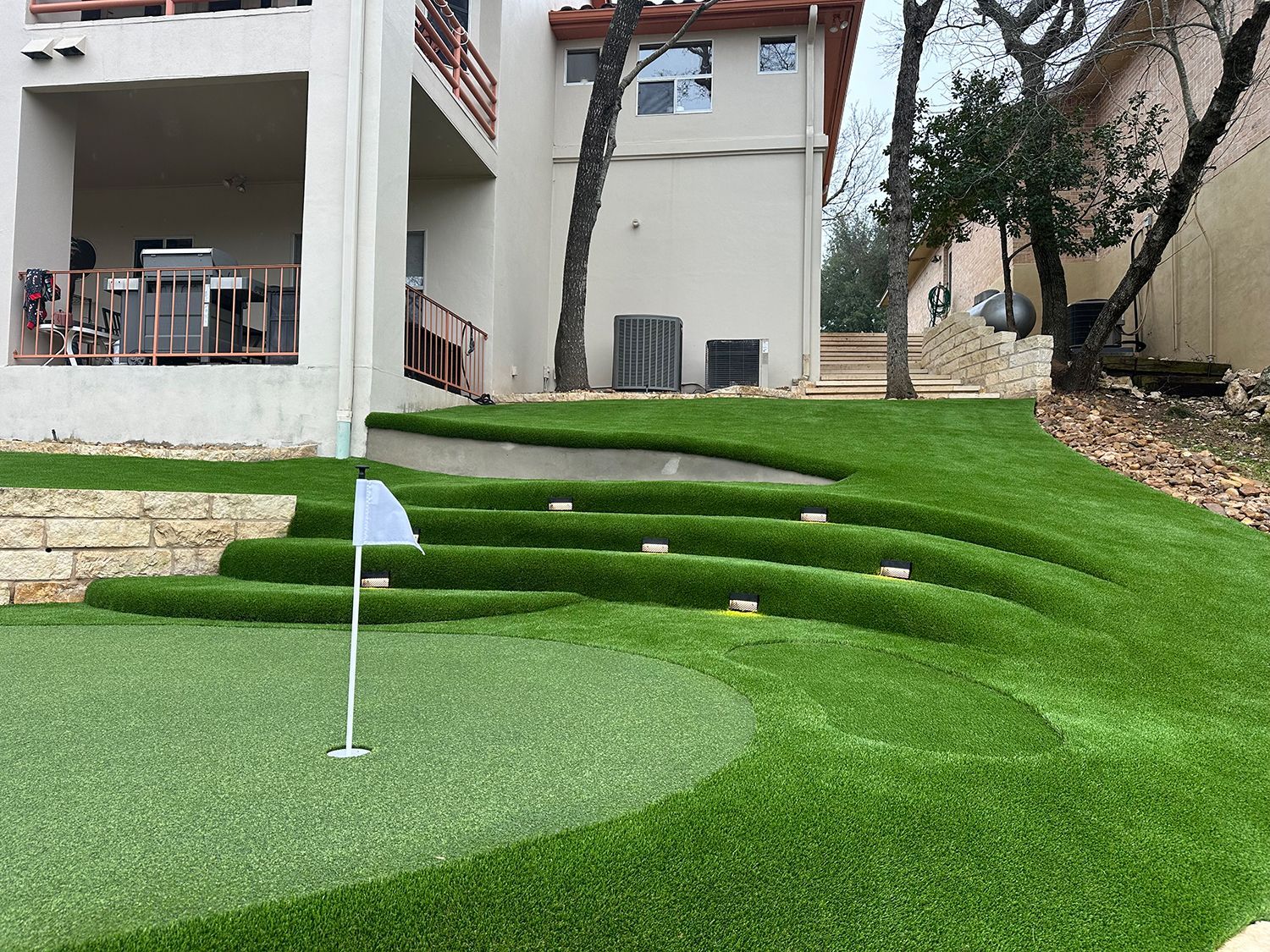 Artificial turf putting green with tiered, landscaped lawn and flag.