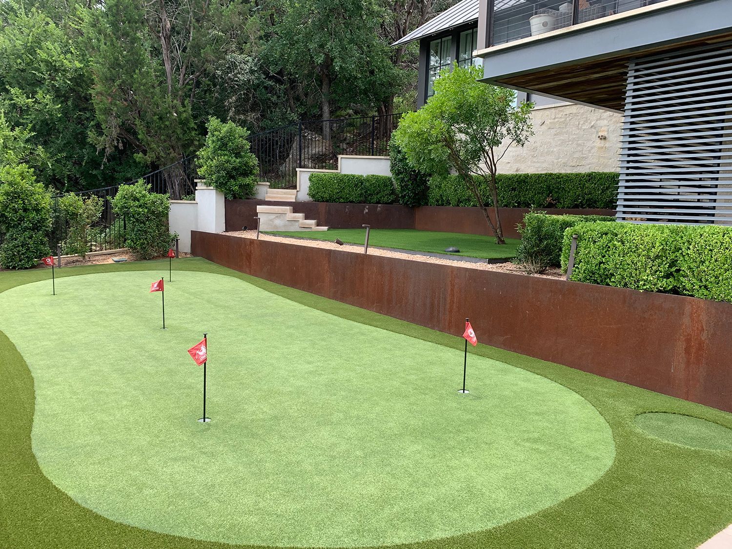 Putting green with red flags, bordered by a rusty-colored retaining wall, lush greenery, and a house in the background.