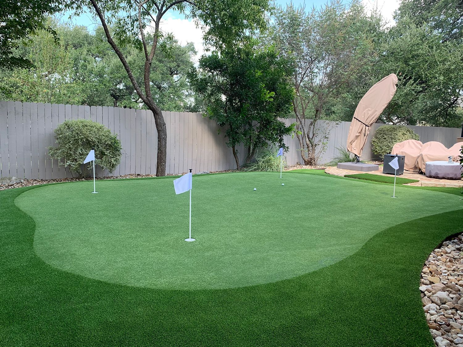 Backyard putting green with white flags and green turf, bordered by rocks and a fence.