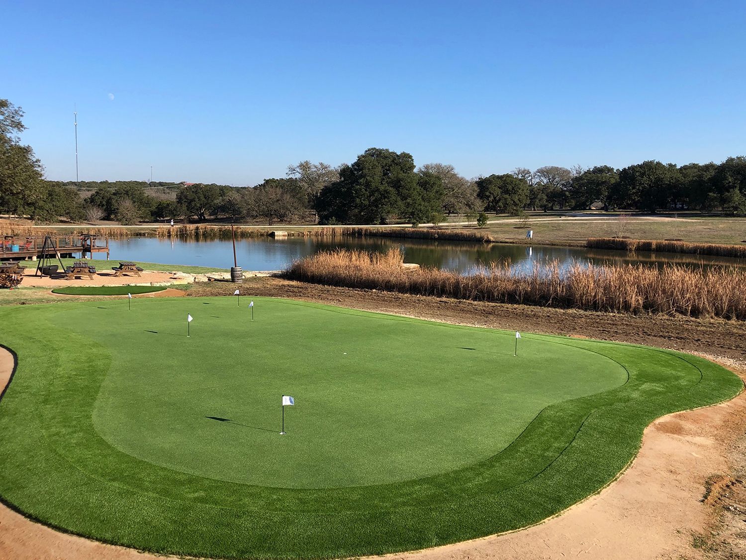 Green golf putting area with flags, pond, and trees on a sunny day.