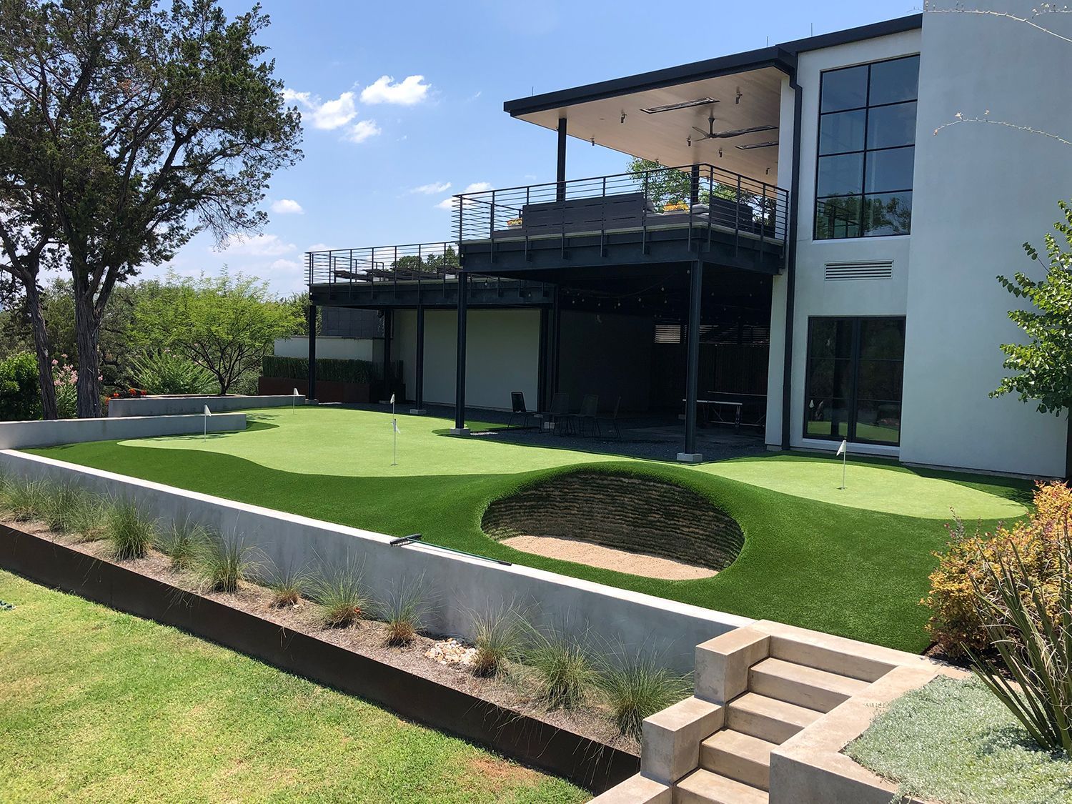 Green putting green with sand trap, next to a modern house with a deck.