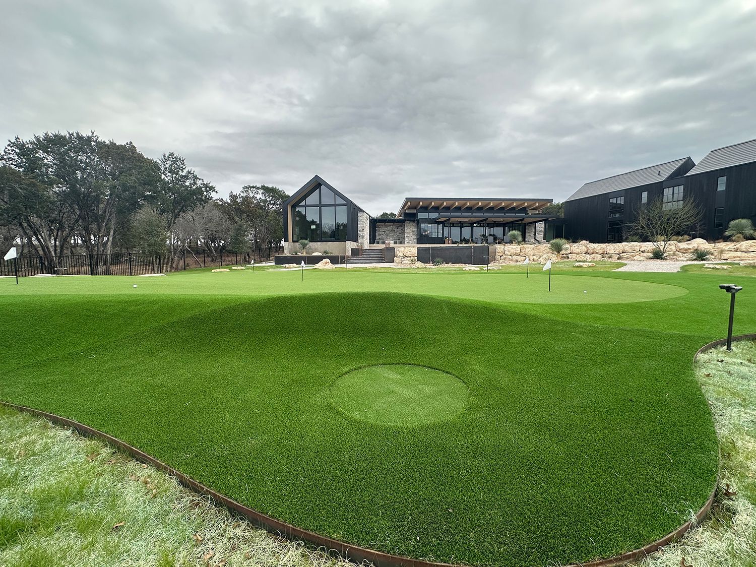 Green artificial turf golf putting green with a modern house in the background under an overcast sky.