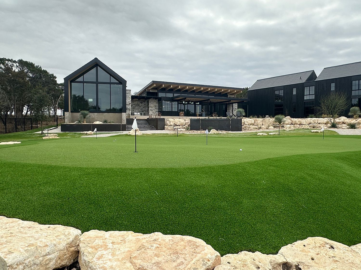 Green putting green with buildings in the background, under cloudy skies.