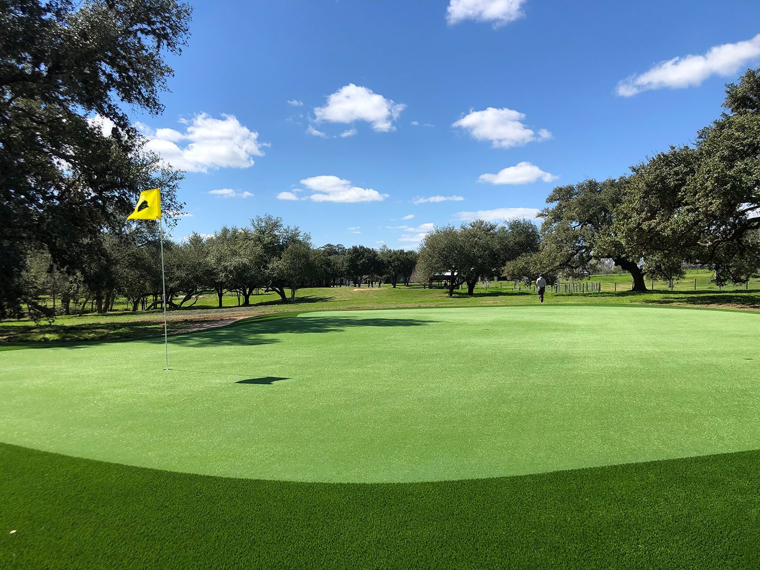 Green golf putting area with a yellow flag, trees, and a person in the distance on a sunny day.