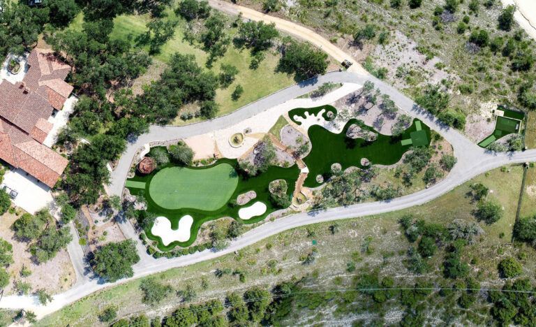 Aerial view of a custom putting green surrounded by a pathway and trees, near a house with a red tile roof.