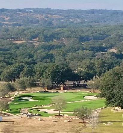Golf course with green fairways and trees, set in a hilly, wooded landscape under a blue sky.