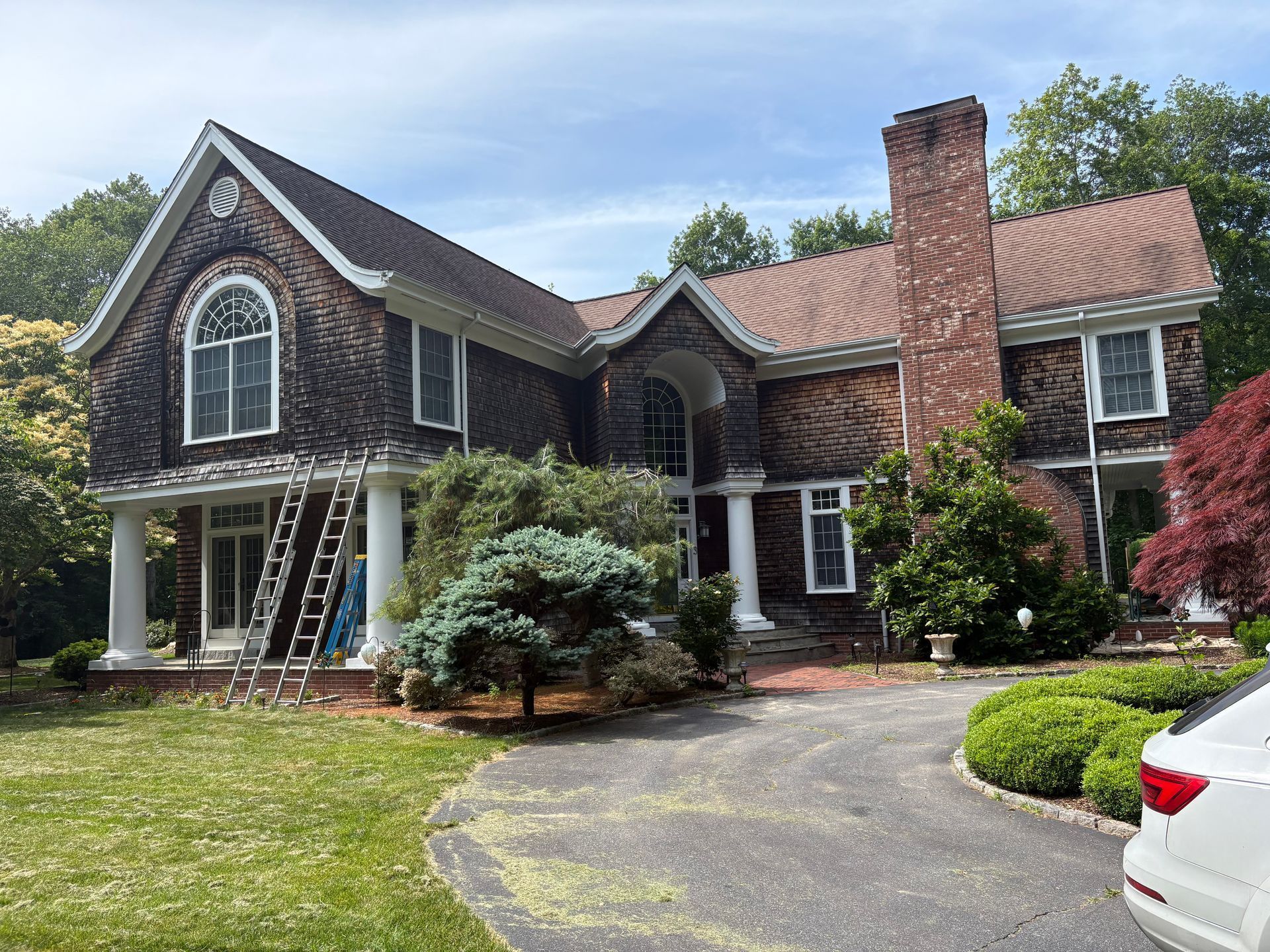 Two-story shingled house with brick chimney and white columns; ladder propped up on the porch.