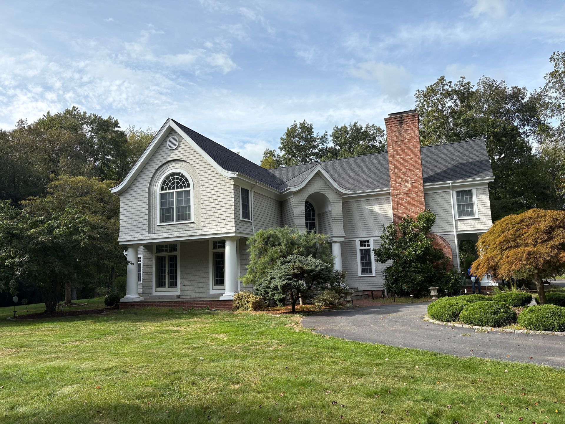 Two-story house with gray siding, white trim, brick chimney, and a curved window, set on a grassy lawn.