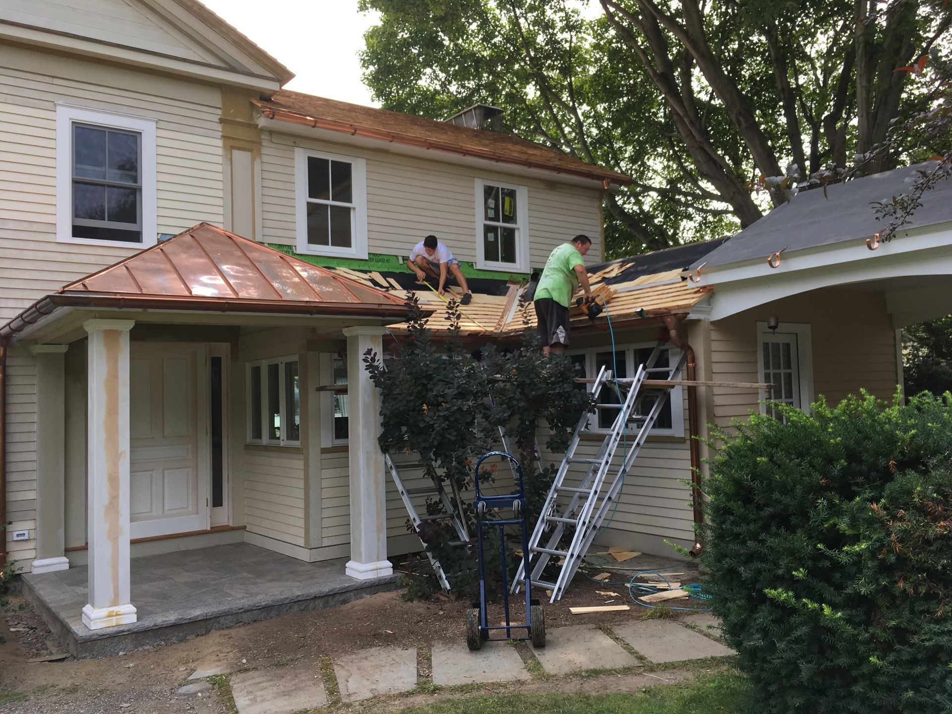 Construction workers on a house roof, removing old shingles. Beige house with copper porch roof.