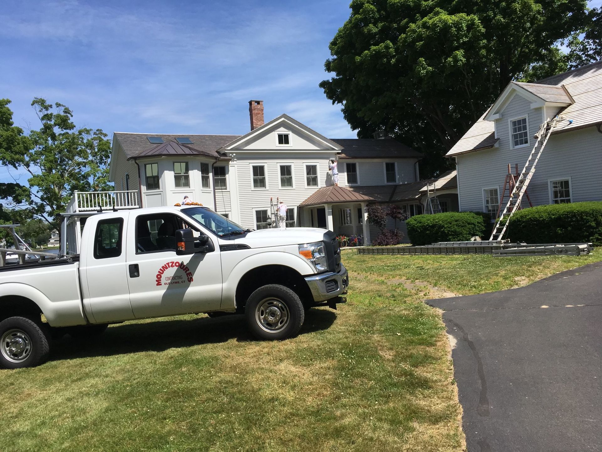 White work truck parked on a grassy lawn in front of a large house being painted.