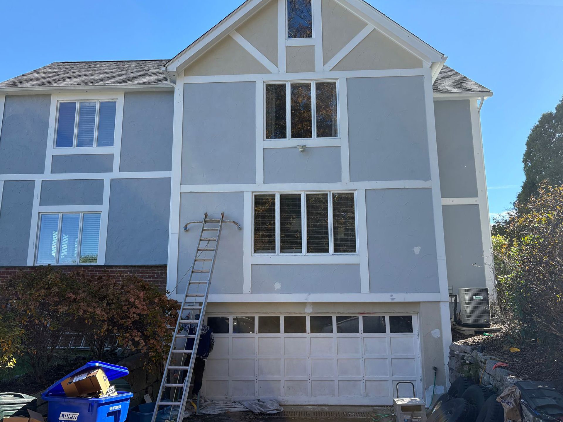 Two-story house with light blue stucco and white trim. Garage door is white. A ladder is leaning against the facade.