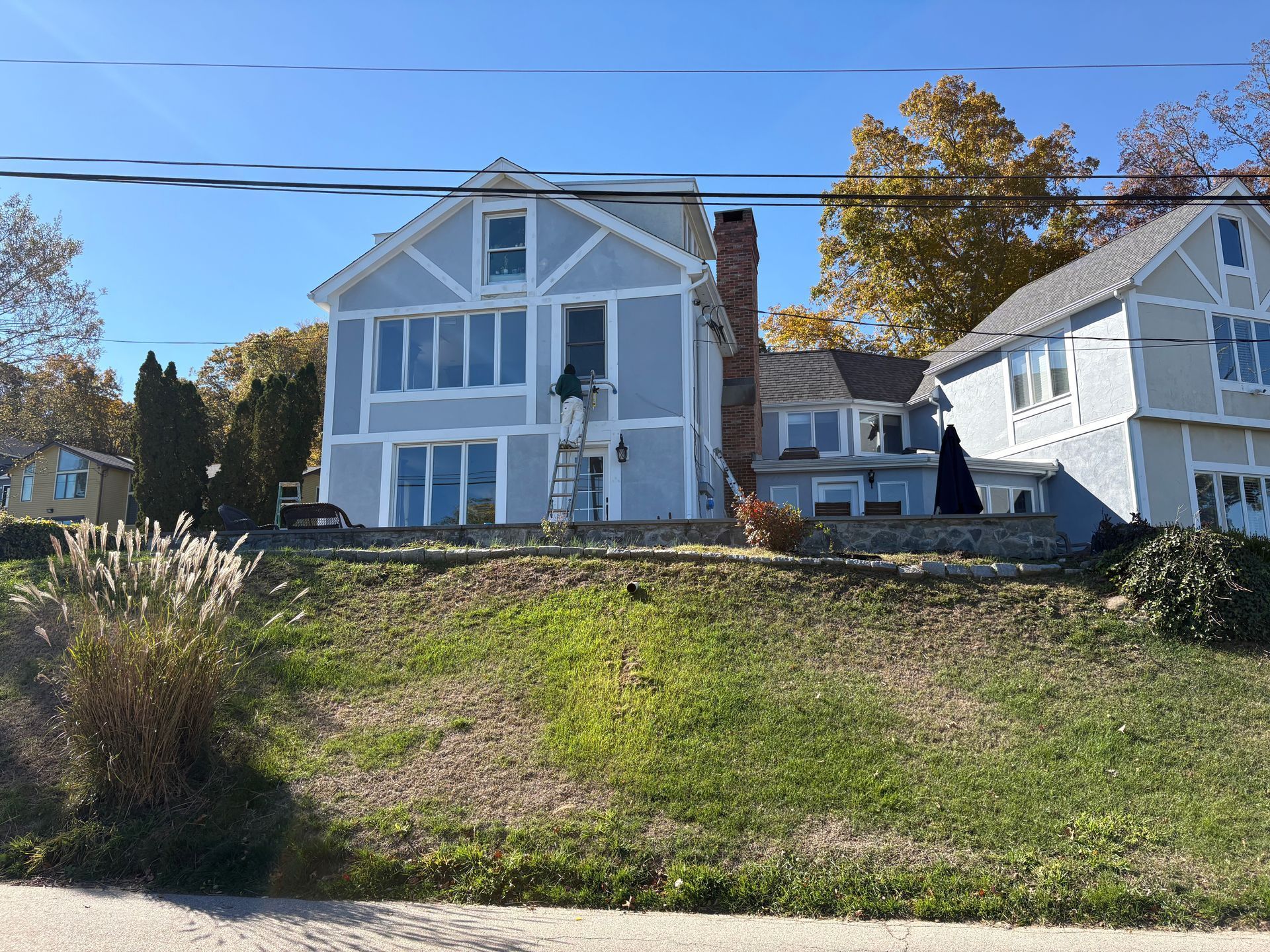 Two-story blue house with large windows, tall brick chimney, and sloped green yard on a sunny day.