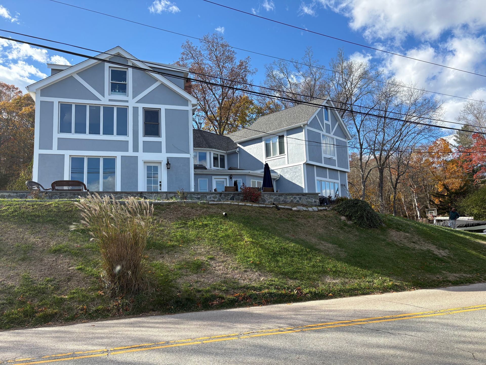 Two-story blue house with white trim on a grassy hill; trees with fall foliage in the background under a blue sky.