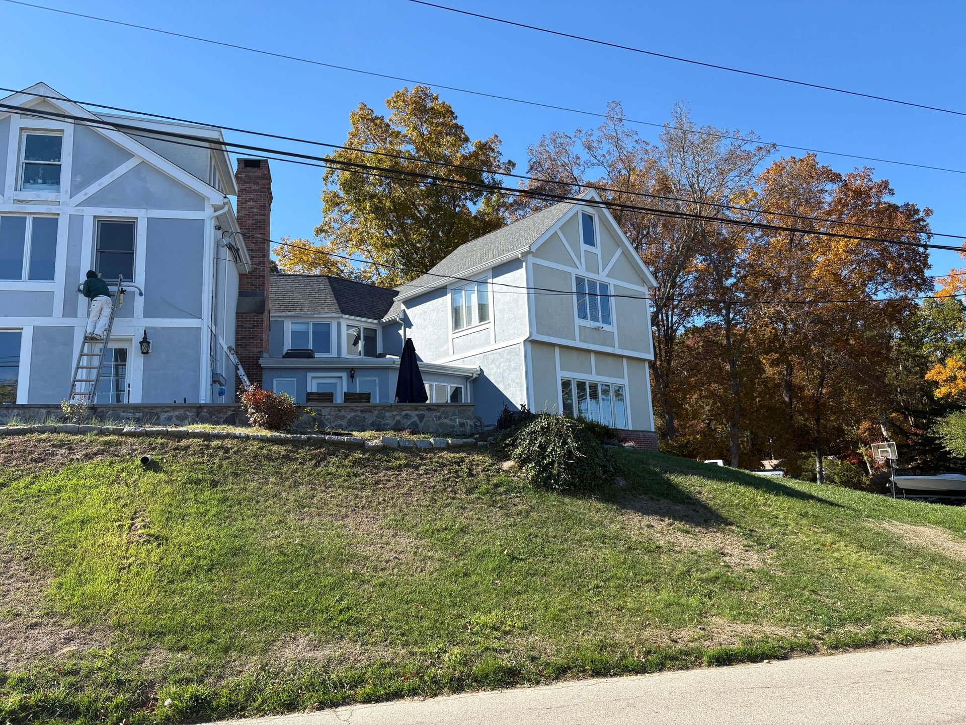 Two-story light blue house with white trim on a grassy hill; fall foliage and clear blue sky.