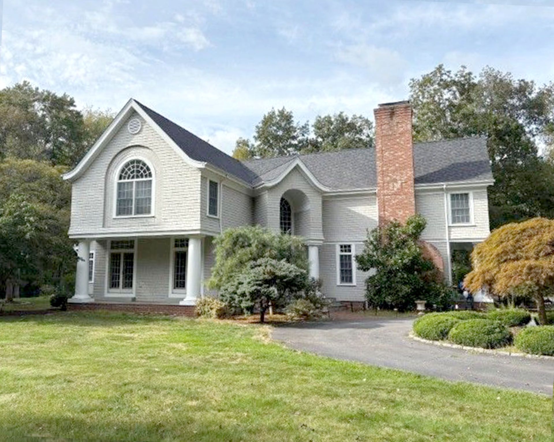 Two-story light gray house with curved architectural features, a brick chimney, and a driveway