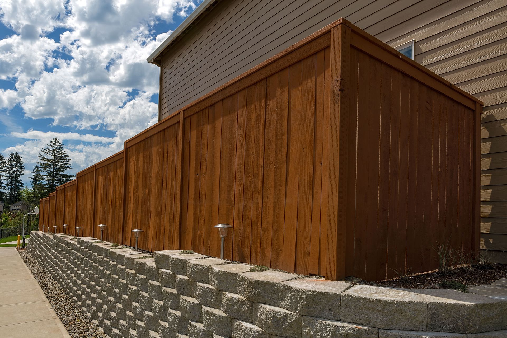 Brown wooden fence on a retaining wall next to a building and sidewalk, under a cloudy sky.