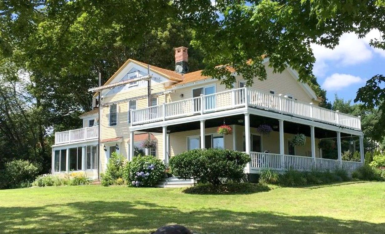 Two-story yellow house with wrap-around porch, white railings, and green lawn, under blue sky