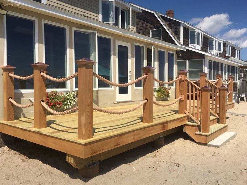 Wooden deck with rope railing on a sandy beach, attached to a light-colored building with large windows.