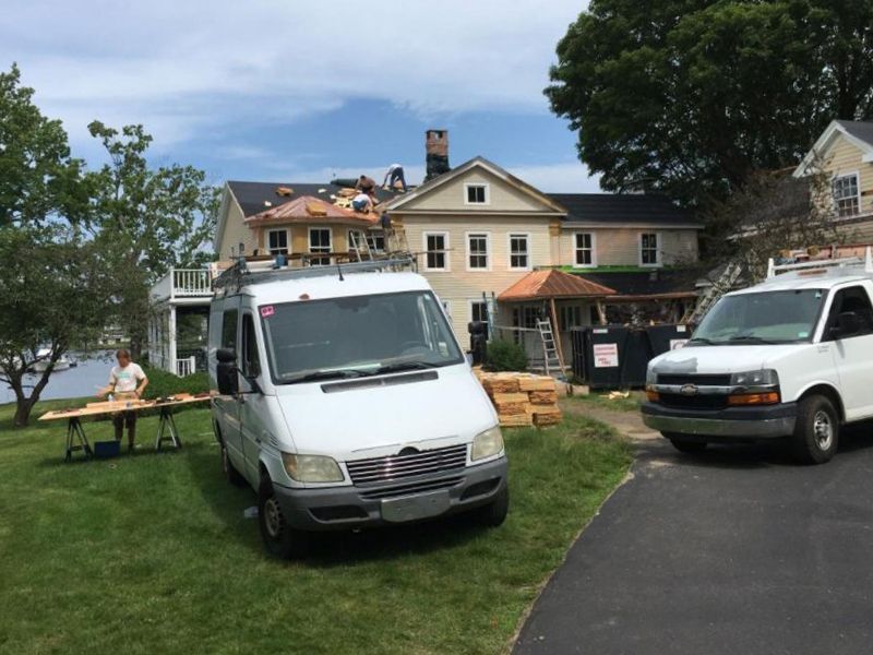 Roofers working on a house roof, with vans parked on the lawn. Sunny day.