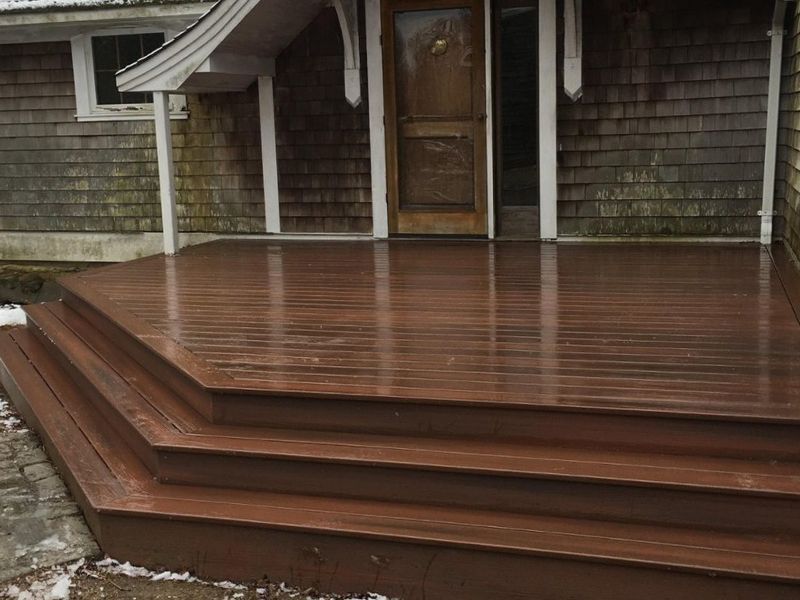 Brown wooden deck and steps leading to a wooden front door, with a snow-covered stone area on the left.