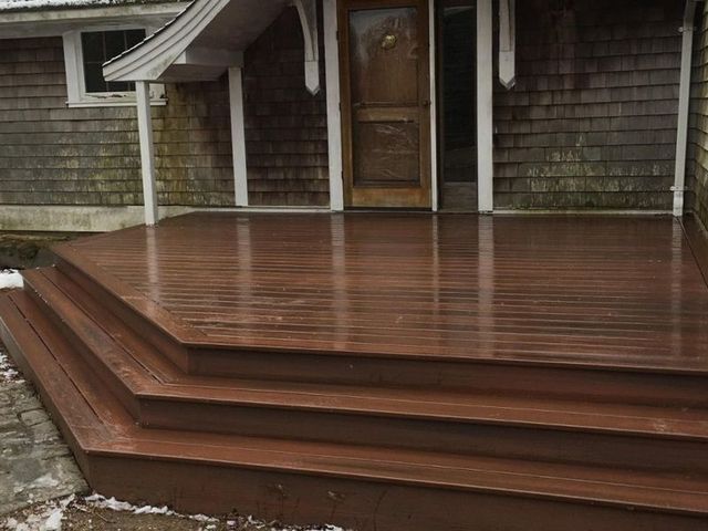 Brown wooden deck and steps leading to a wooden front door, with a snow-covered stone area on the left.