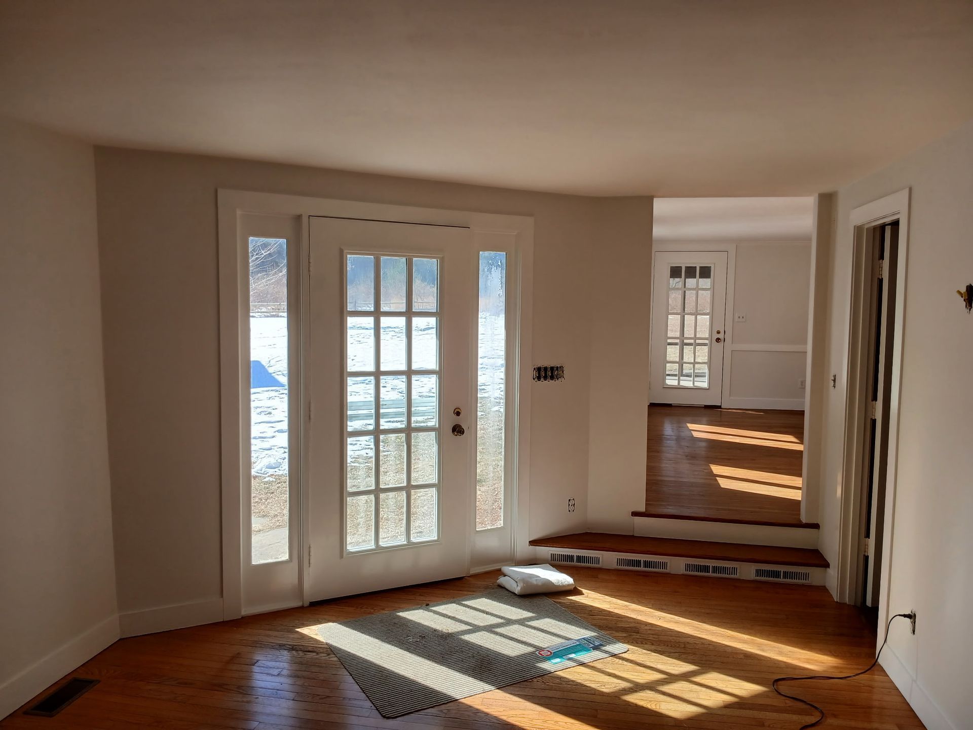 Interior room with French doors and wood flooring. Sunlit, leading to another room.