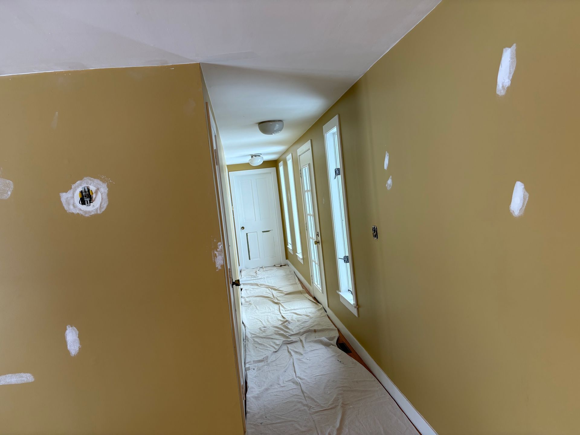 Yellow hallway with patched walls, leading to a closed white door and windows.