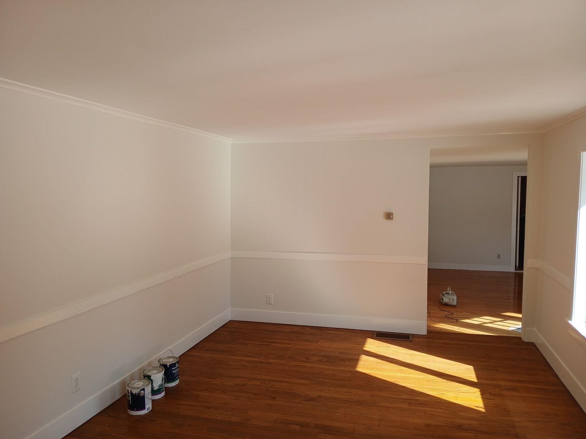 Empty room with white walls, wainscoting, and hardwood floors; paint cans on the floor.